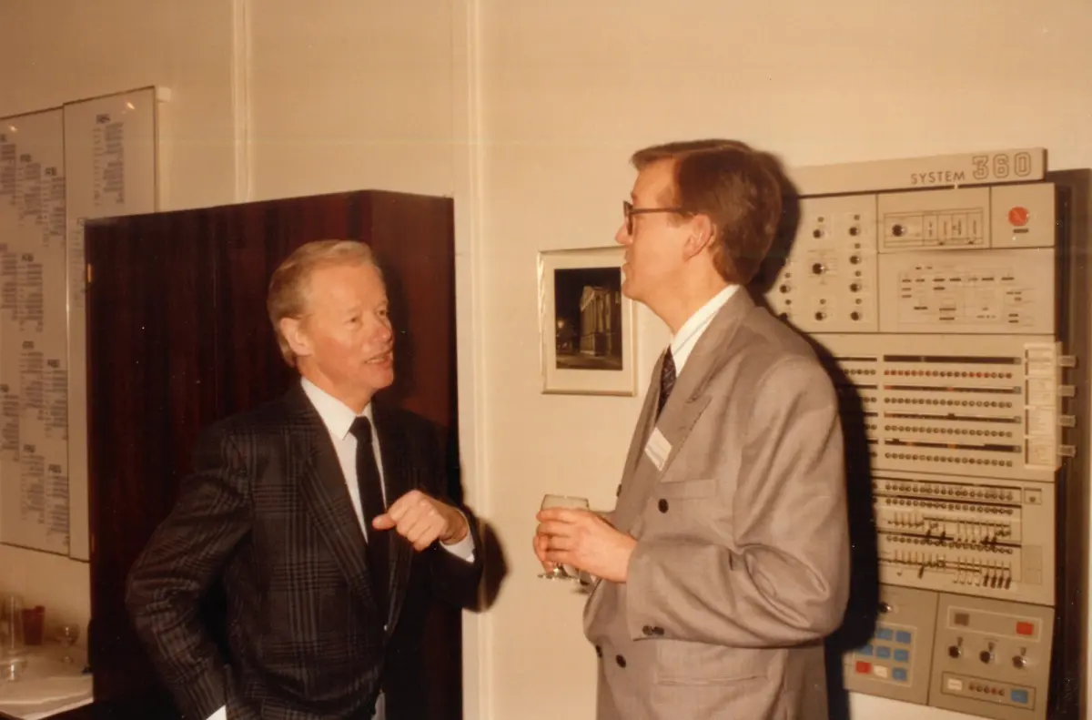 Vagn Sølvkjær in a dark checked suit talking with another man, a large computer control panel on the wall behind them