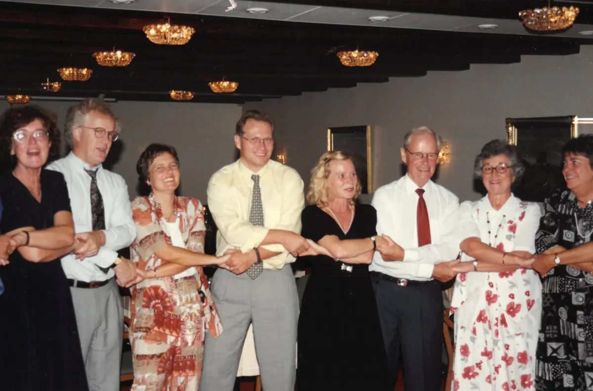 Eight people in a row hold hands with crossed arms under crystal chandeliers, smiling