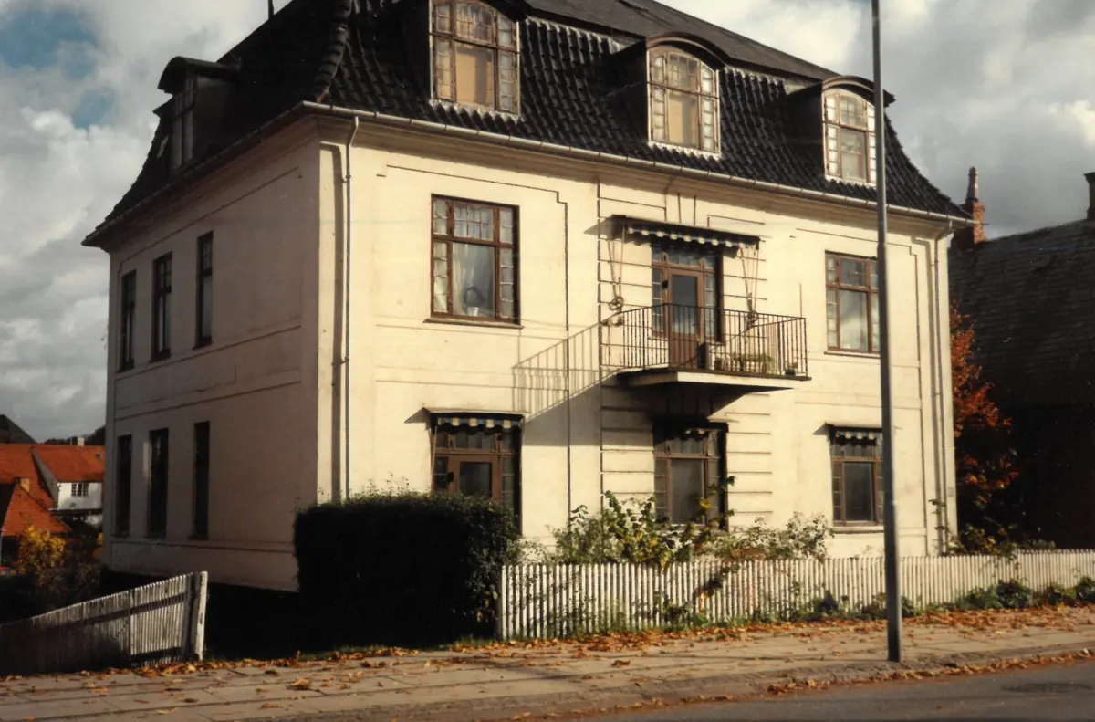 White two-storey house with a dark mansard roof, dormer windows and a balcony, behind a white picket fence