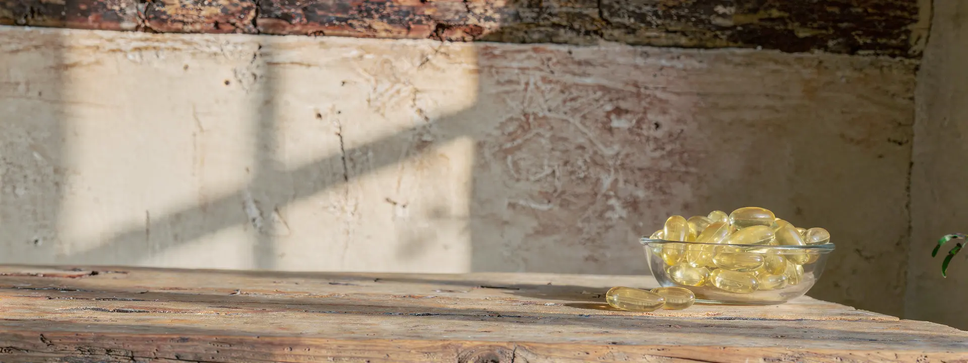 A glass bowl of pale yellow oblong softgels on a rustic oak table in warm sunlight
