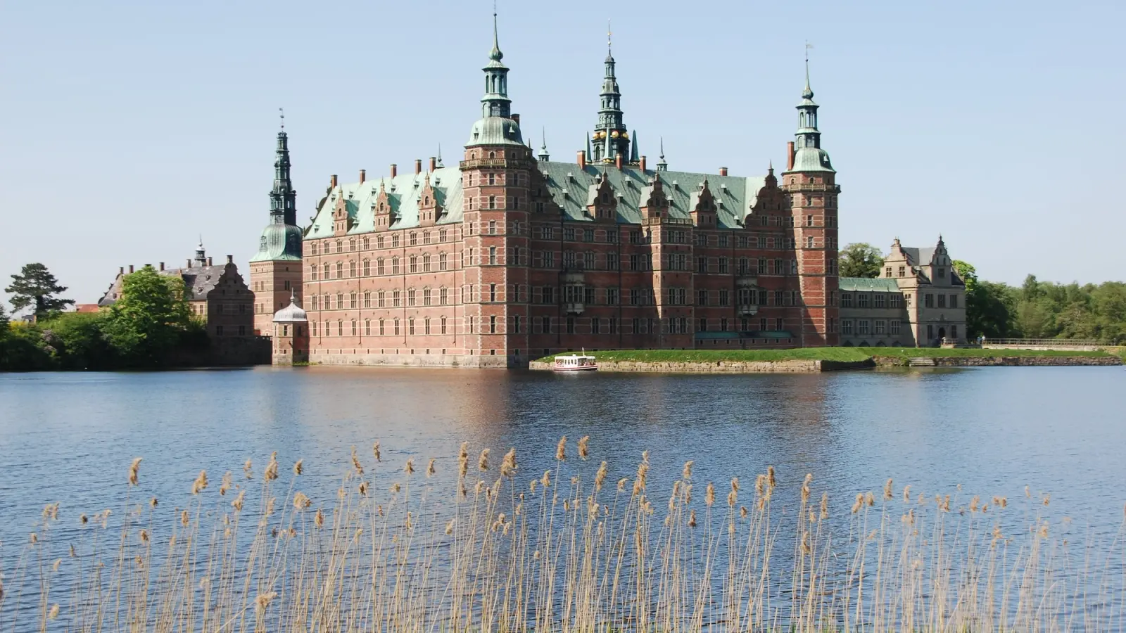 Frederiksborg Castle’s red-brick towers and green copper roofs across the castle lake, with reeds in front